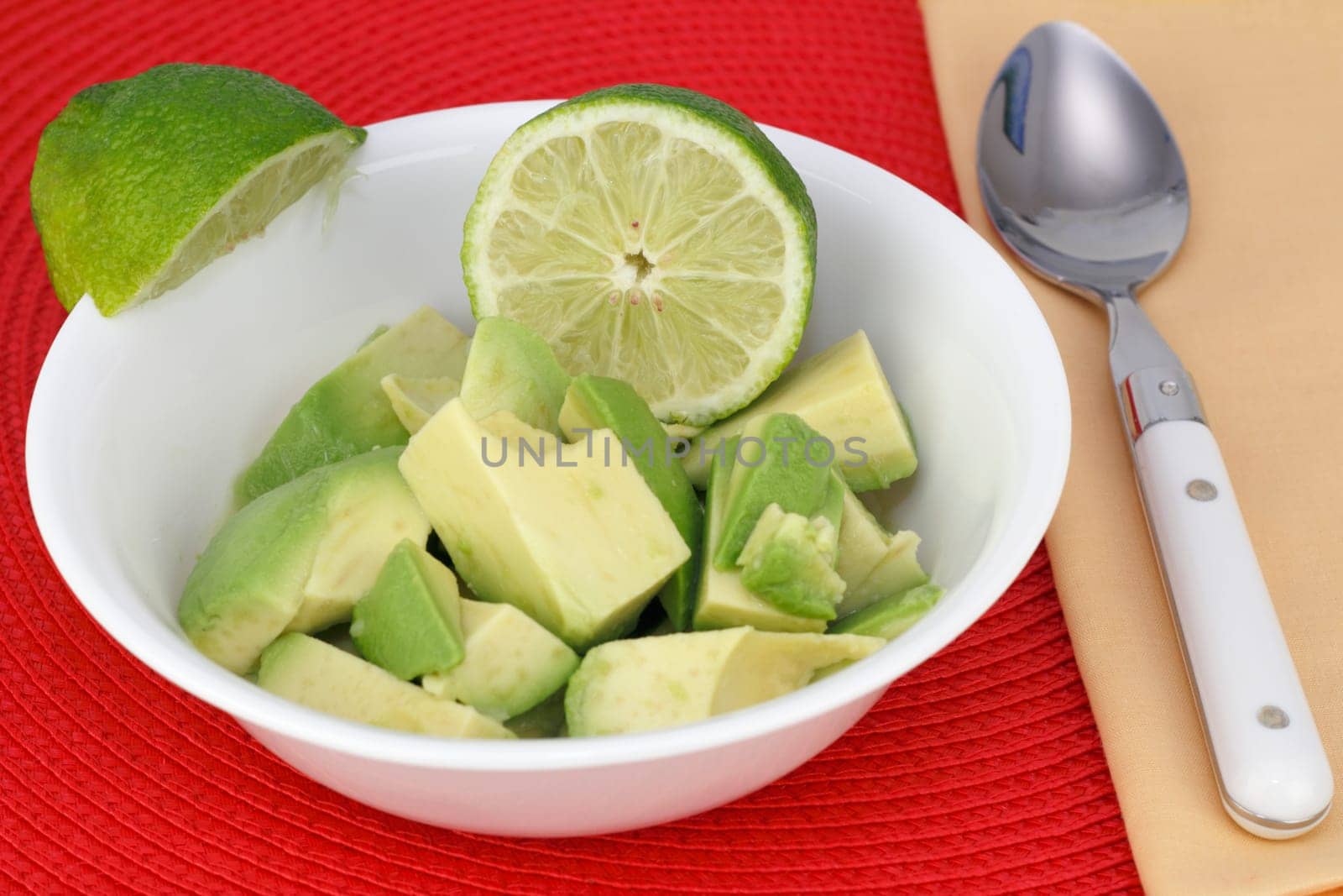 Avocado pieces in a white bowl with lime juice and lime wedges on the side of the bowl. A spoon is near by on a yellow napkin and everything is on a red placemat.