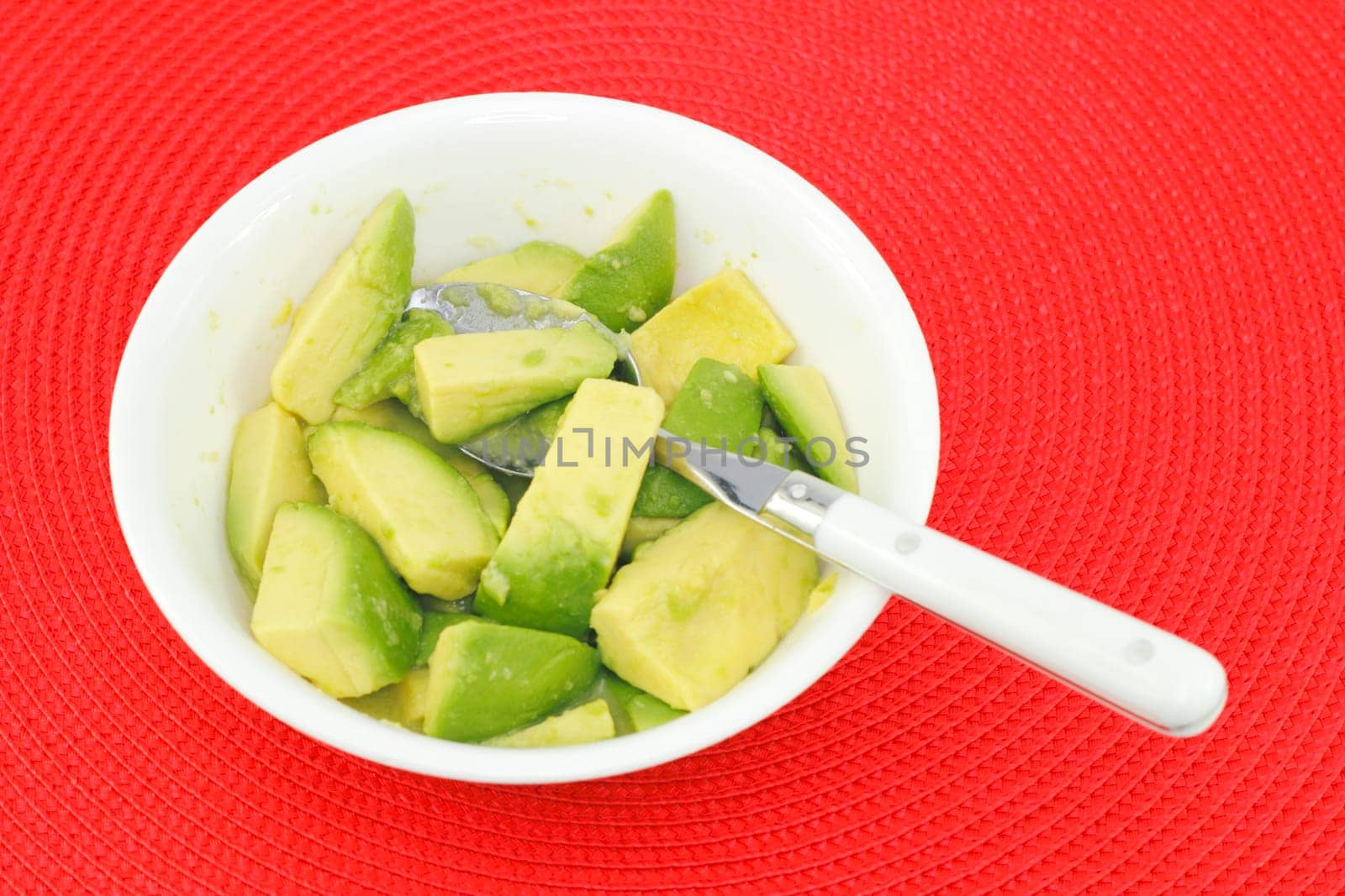 Close up of a spoon in a white bowl of just avocado pieces mixed with lime juice on a red placemat.