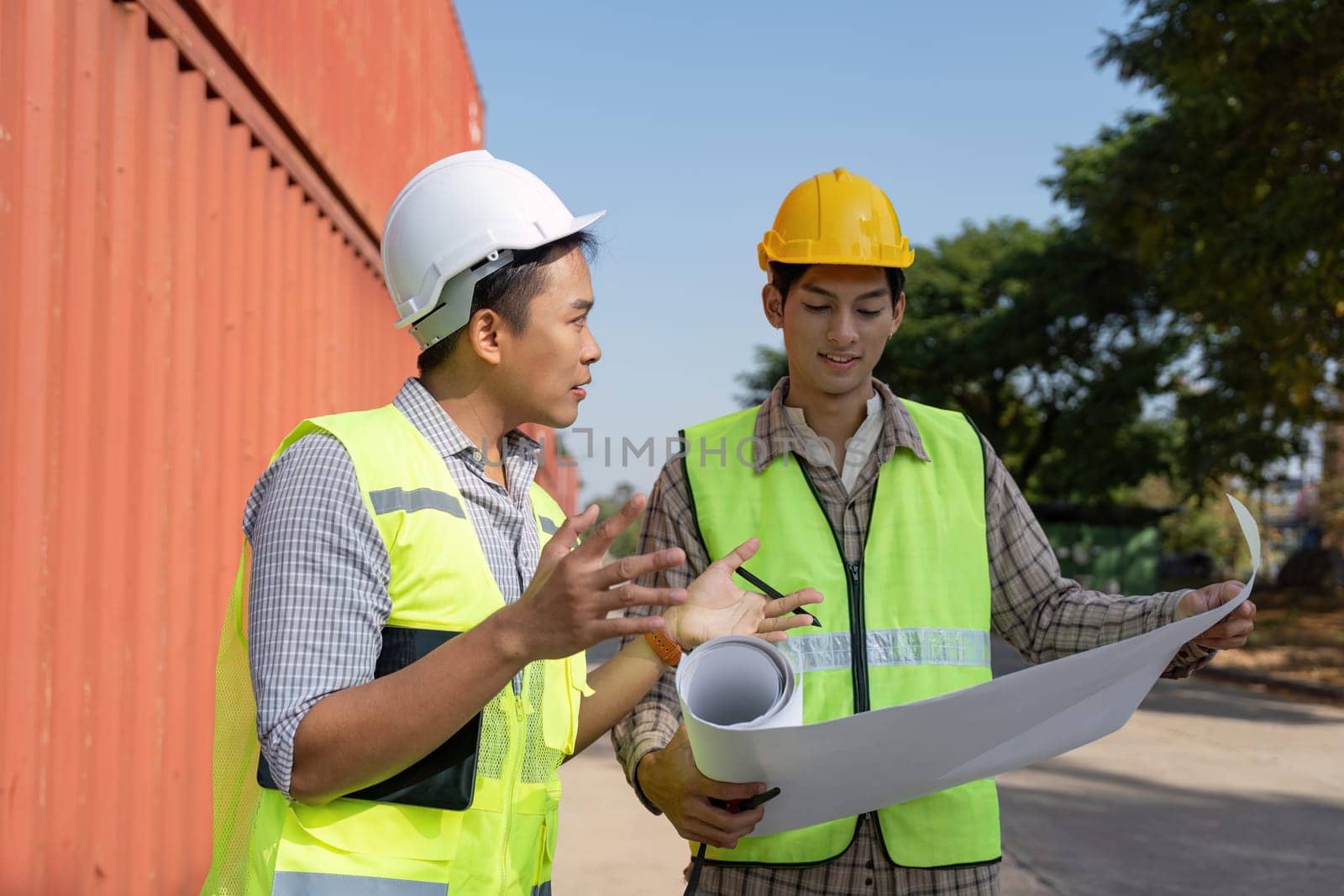 Two engineers work together on logistics strategies, examining blueprints in a shipping container yard.
