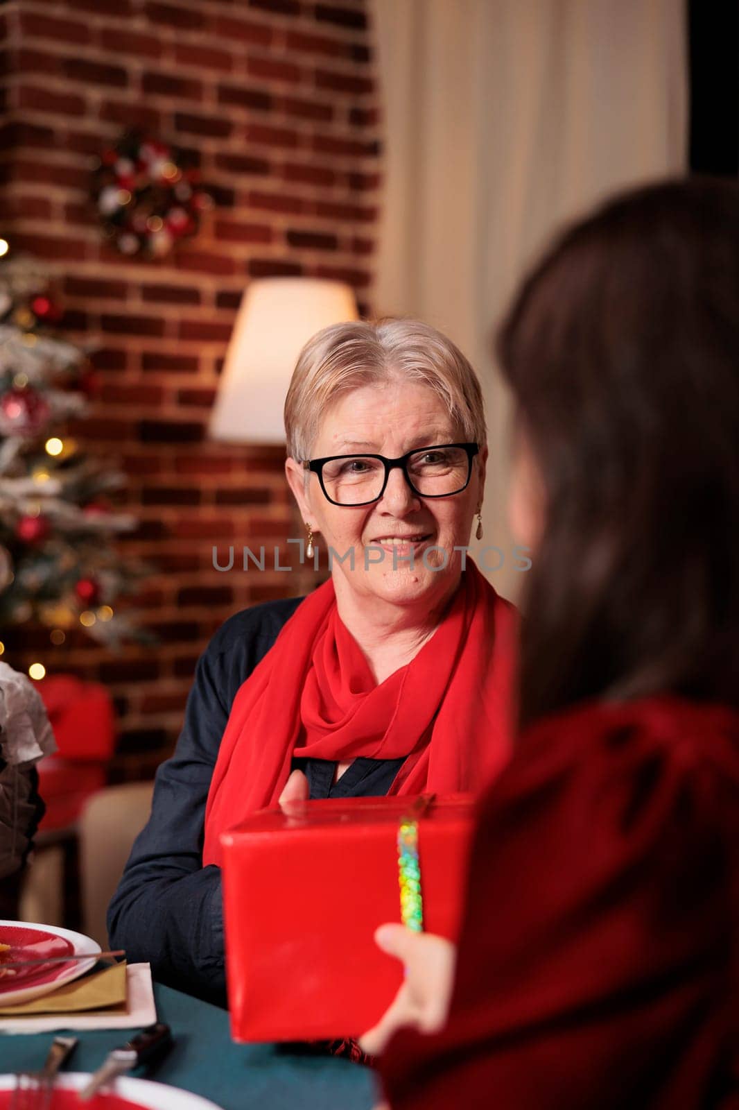 Woman giving present to her mother at christmas dinner celebration, feeling cheerful during december celebration. Family exchanging gifts and enjoying good time at home with people.