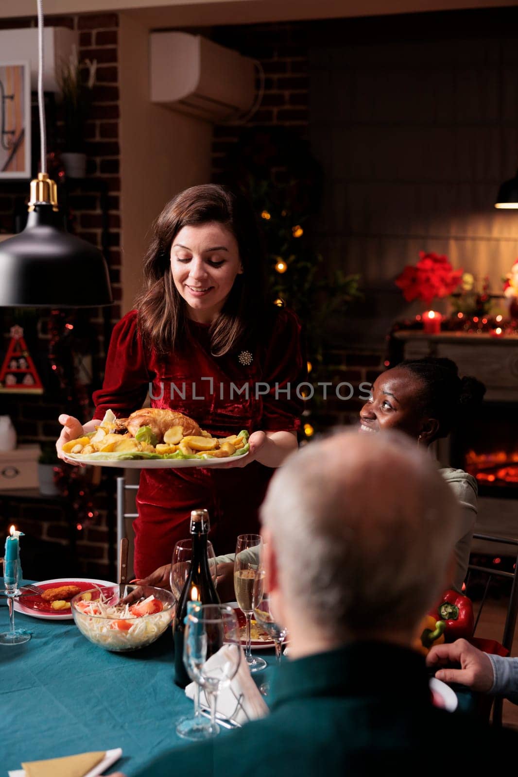 Woman putting festive food on table, enjoying christmas dinner and seasonal celebration with diverse friends and family at home. People feeling happy eating homemade meal and drinking alcohol.