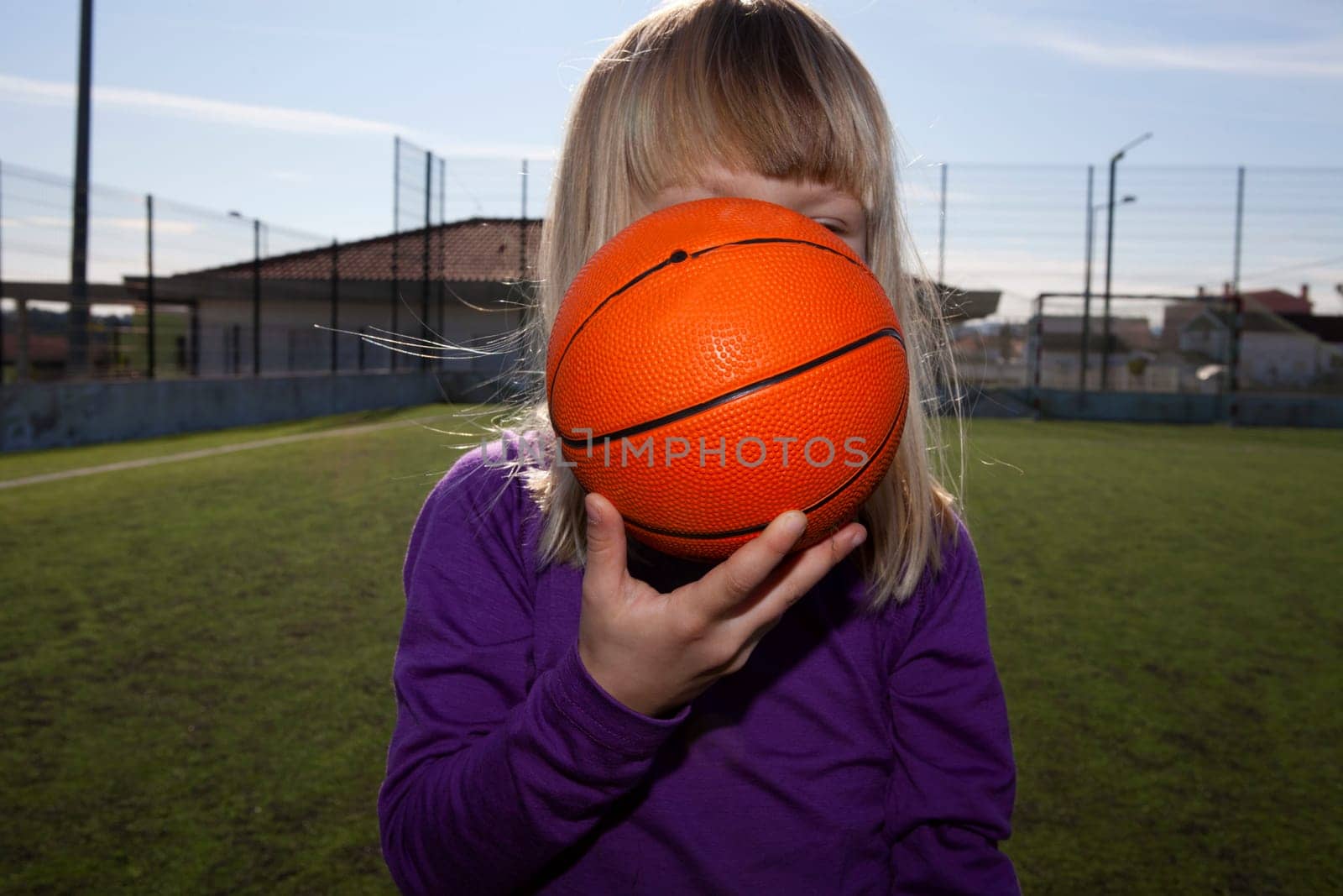 Little girl standing and hiding behind a basketball on an outdoor court