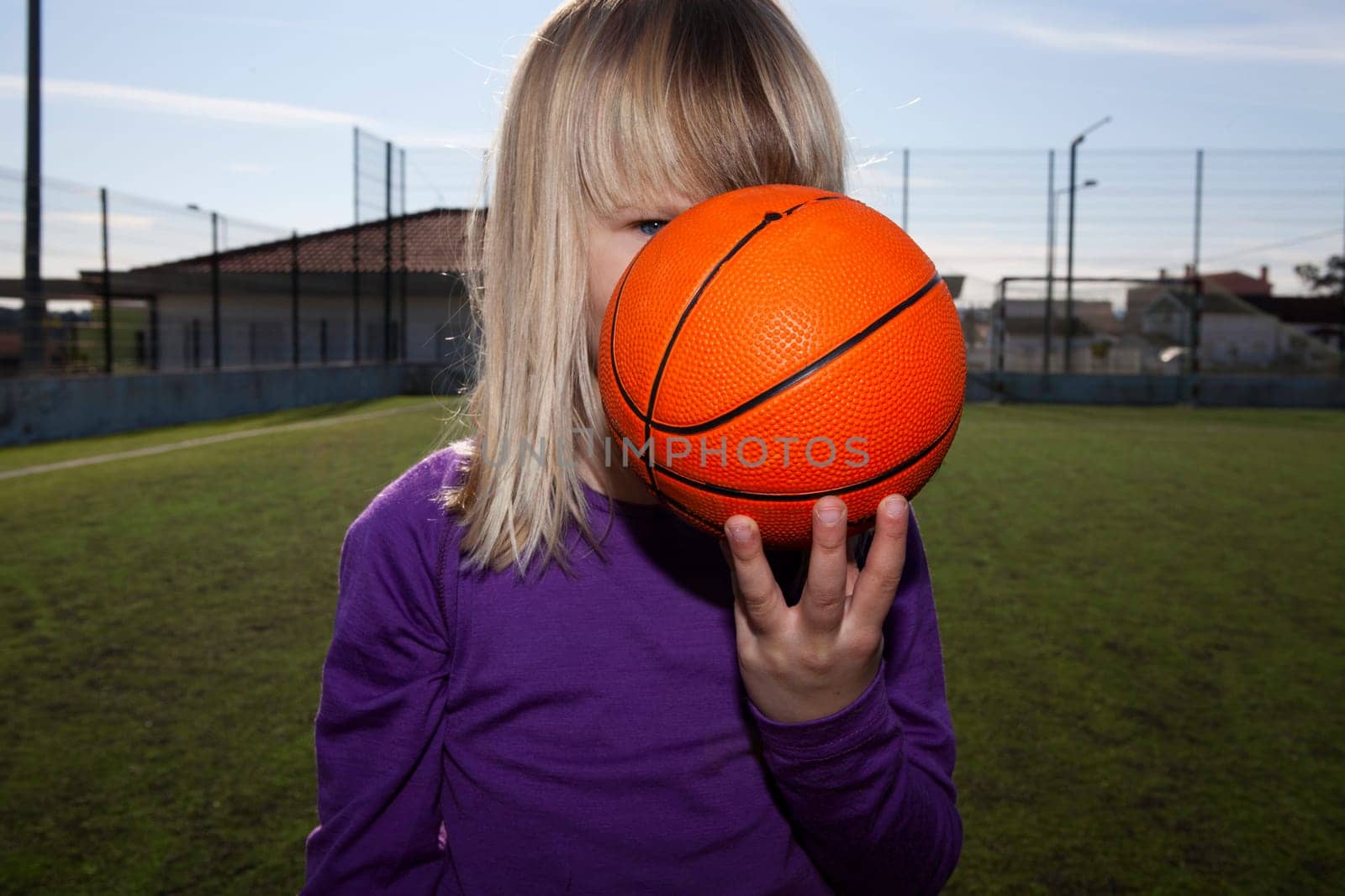 Little girl standing and hiding behind a basketball on an outdoor court