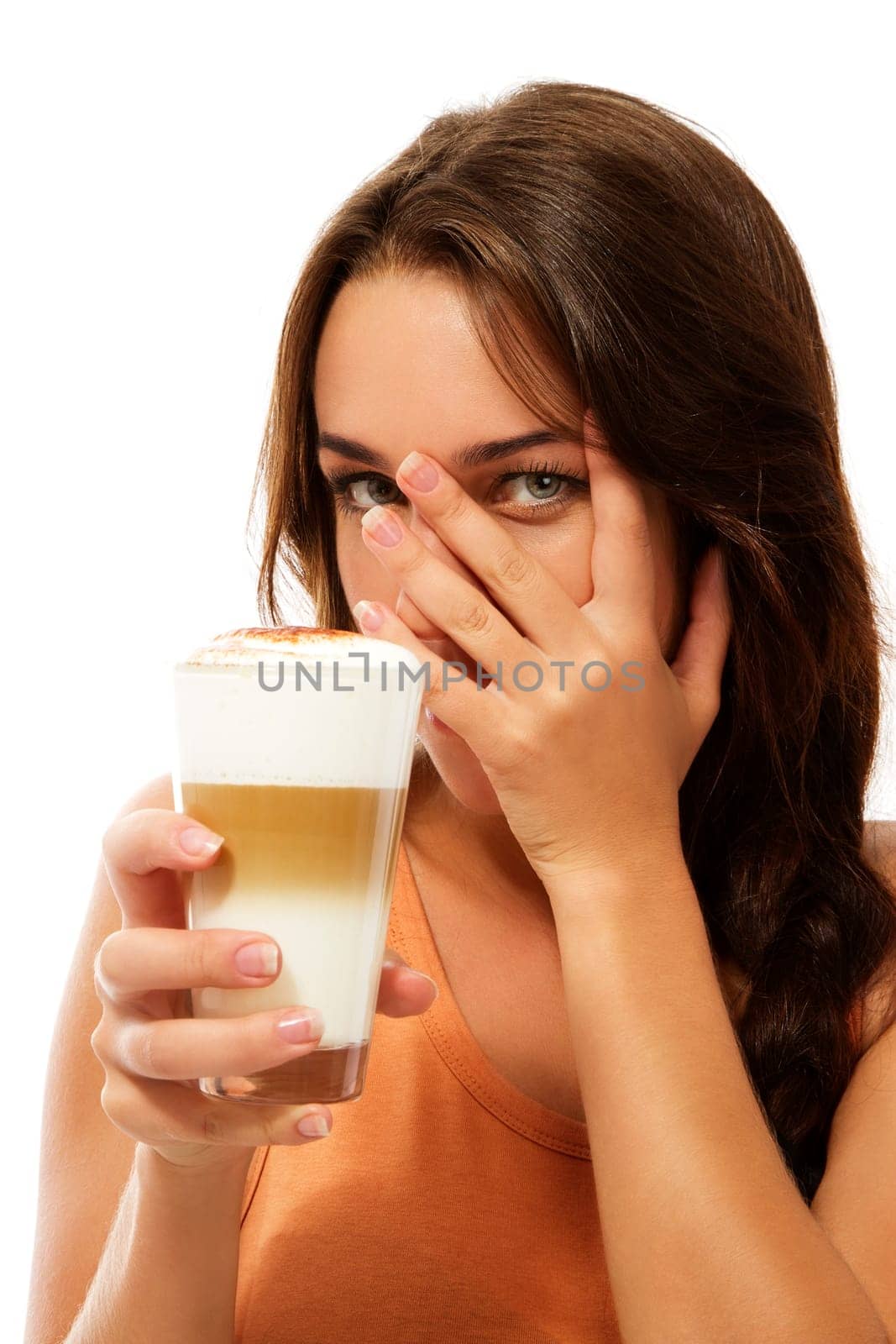Young woman with a glass of latte macchiato hides her face behind her hand against white background