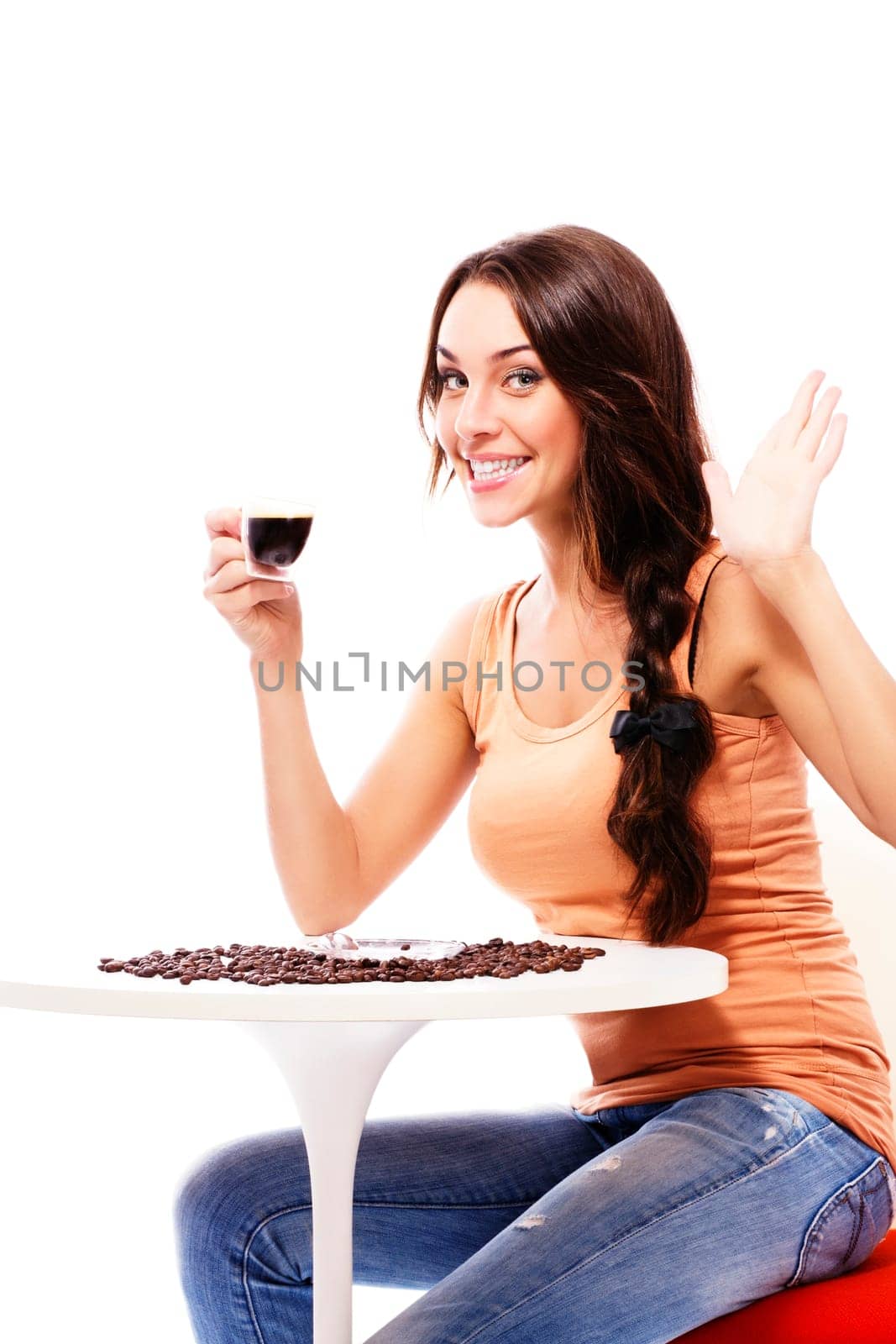 waving laughing young woman with a cup of espresso coffee sitting at a table against a white background