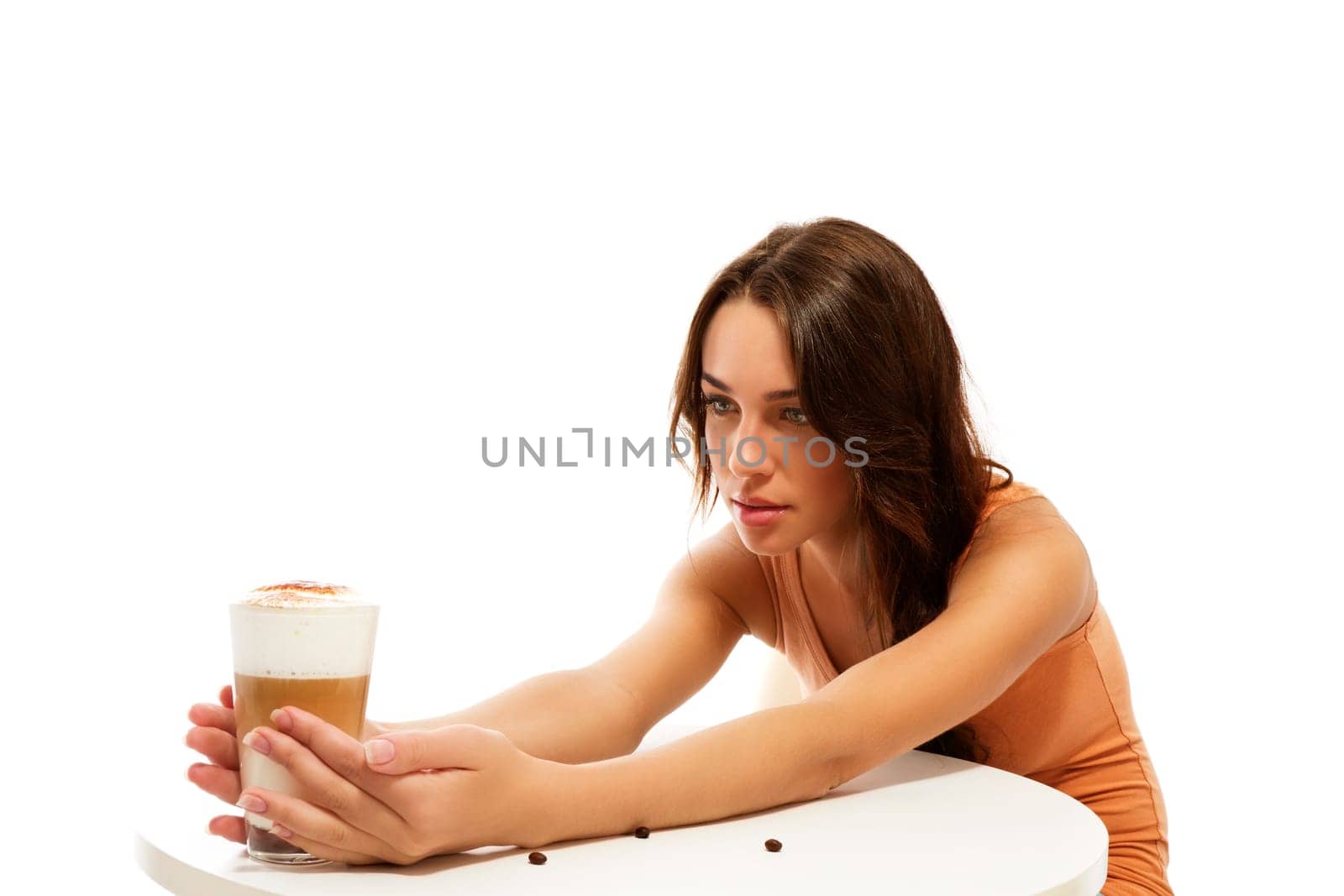 Young woman looking at the glass of latte macchiato coffee on the other side of the table against white background