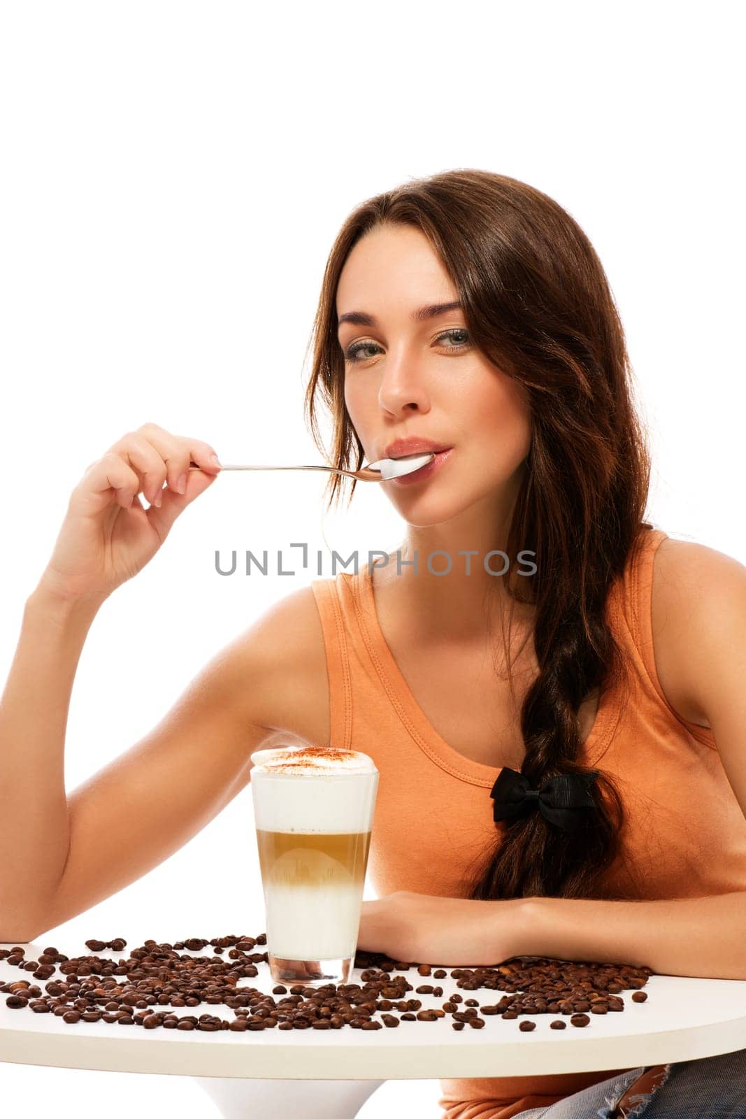 Young woman sitting at a table with a glass of latte macchiato coffee with spoon in mouth against white background