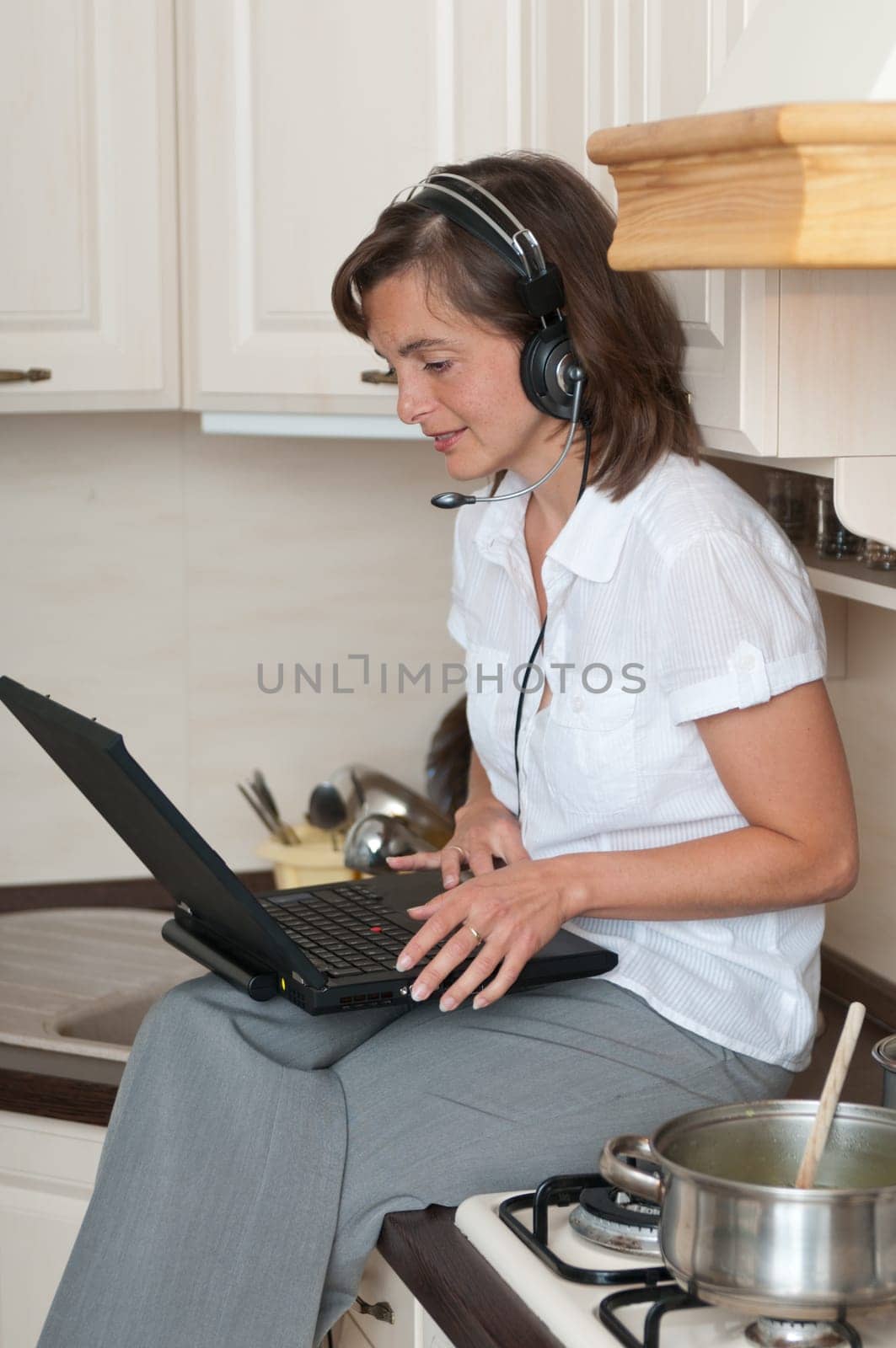 Young business person with headset working from home on laptop while cooking meal in kitchen