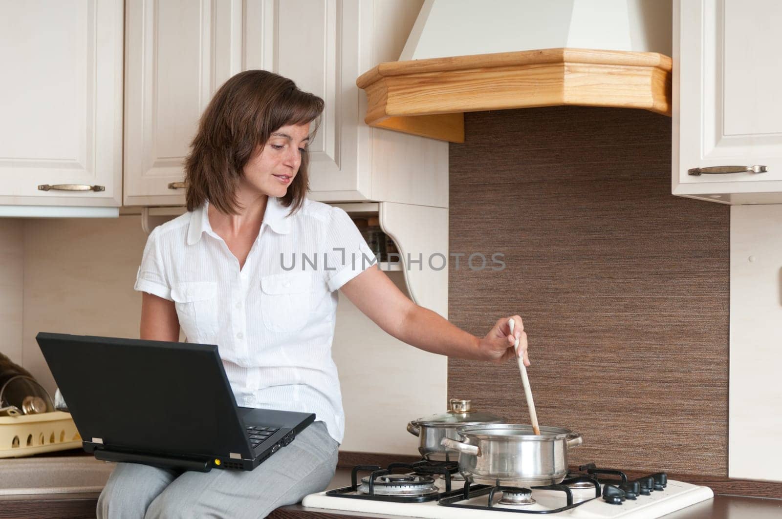 Young woman cooking meal in kitchen according to receipt from laptop (internet)