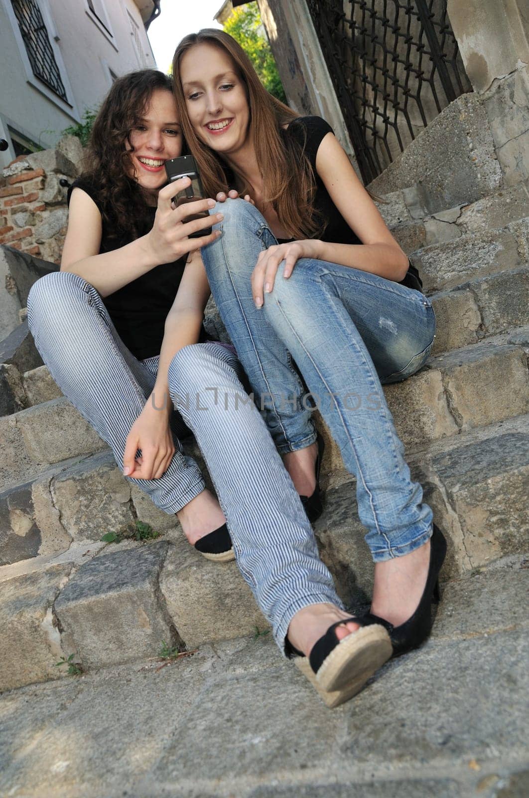 Youth lifestyle - two smiling friends (girls) siting on stairs outdoors with mobile phone