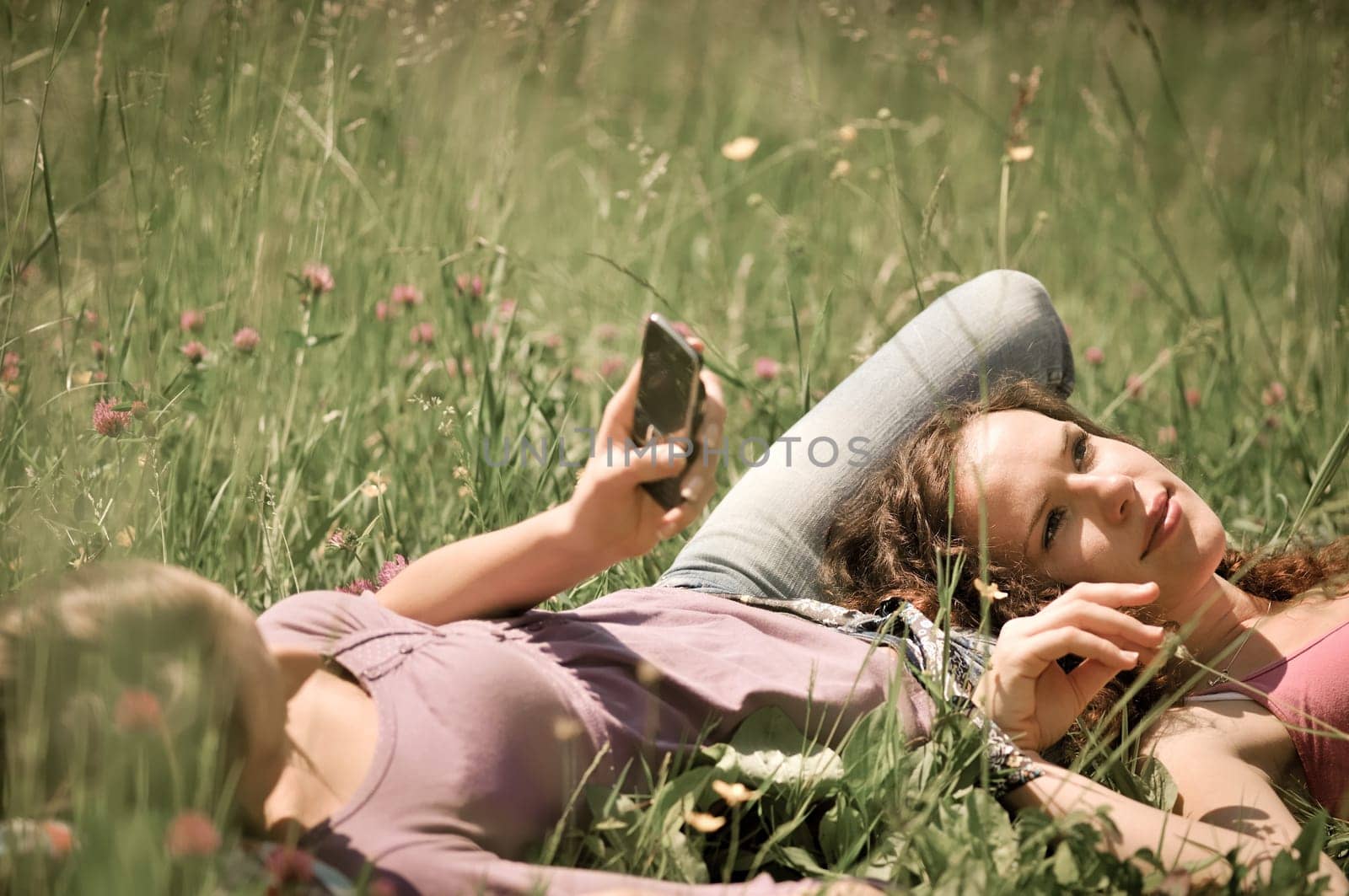 Two friends (teenage girls) relax lying on grass outside in nature - one girl is using cellphone