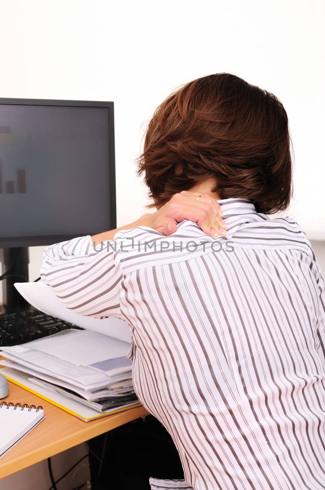 Business woman with neck pain. Focus on hand on neck with blurred monitor and documents on table.