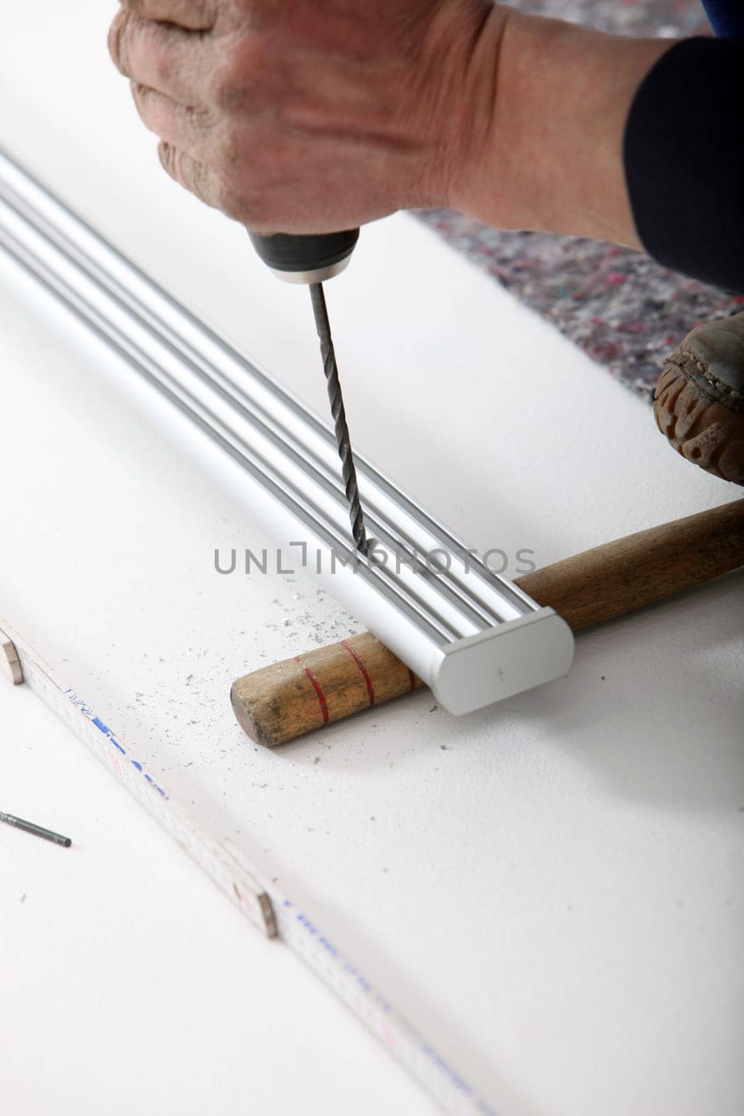 An artisan&#039 s hand using a portable cordless drill to drill a hole in a length of aluminium track supported at ground level.