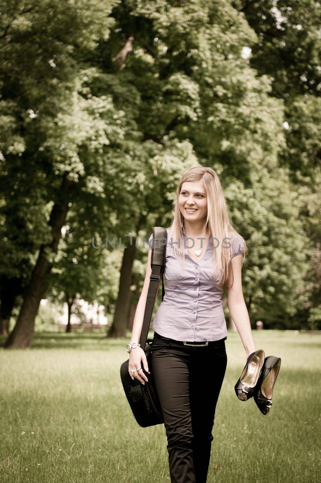 Young business woman wit laptob bag walking outdoors in park and holding shoes