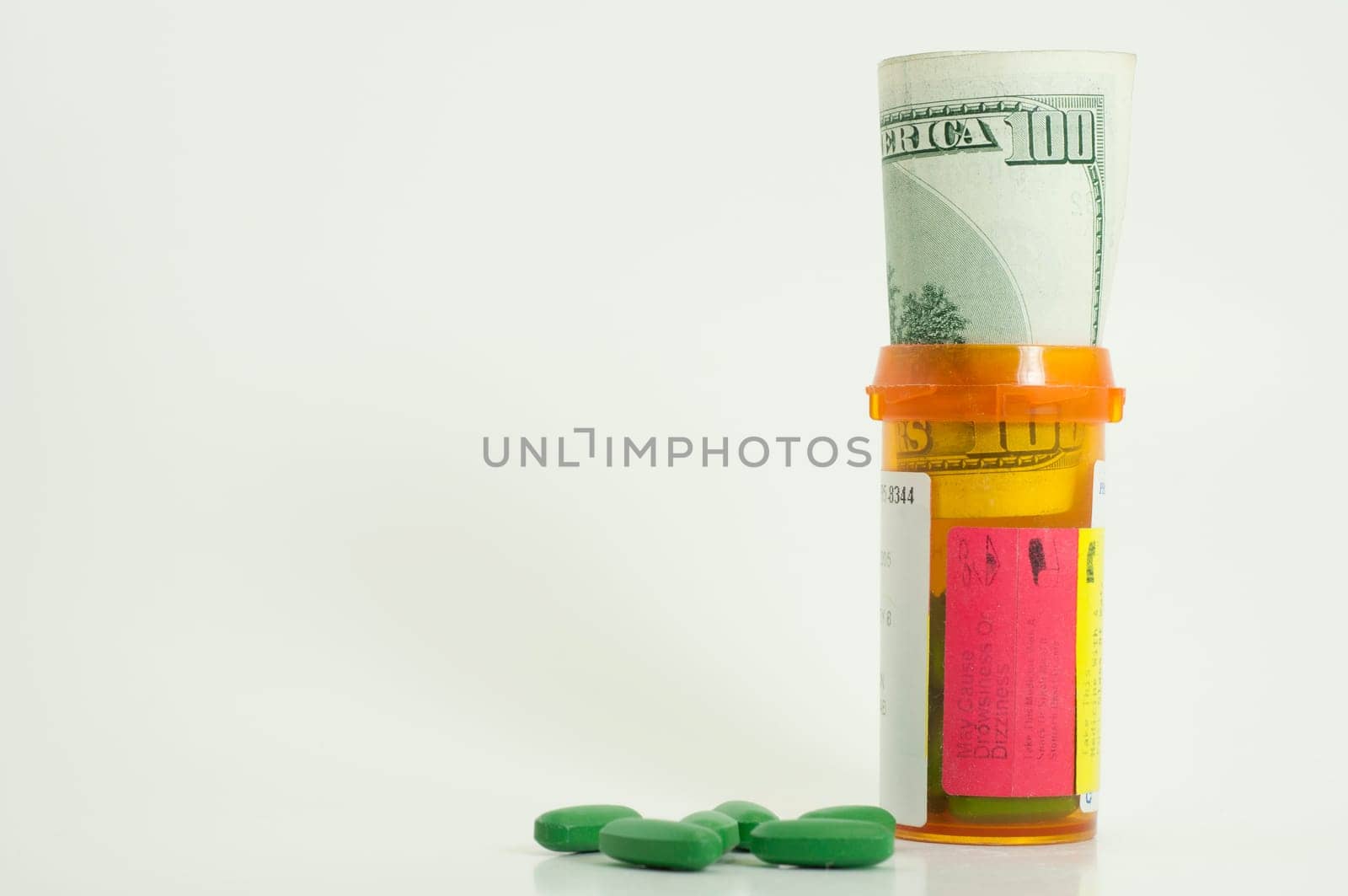 prescription bottle with one hundred dollar bill inside and green pills spilled on white background