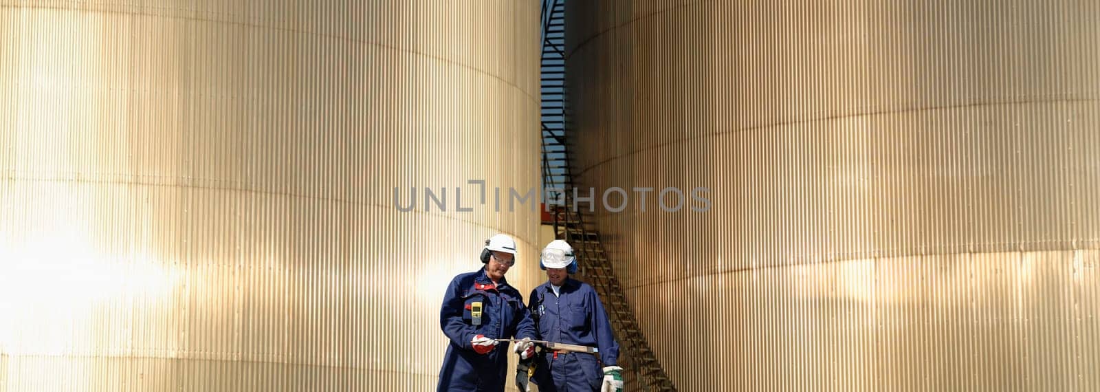 two oil and gas workers, large shiney fuel-storage towers in background, panoramic view