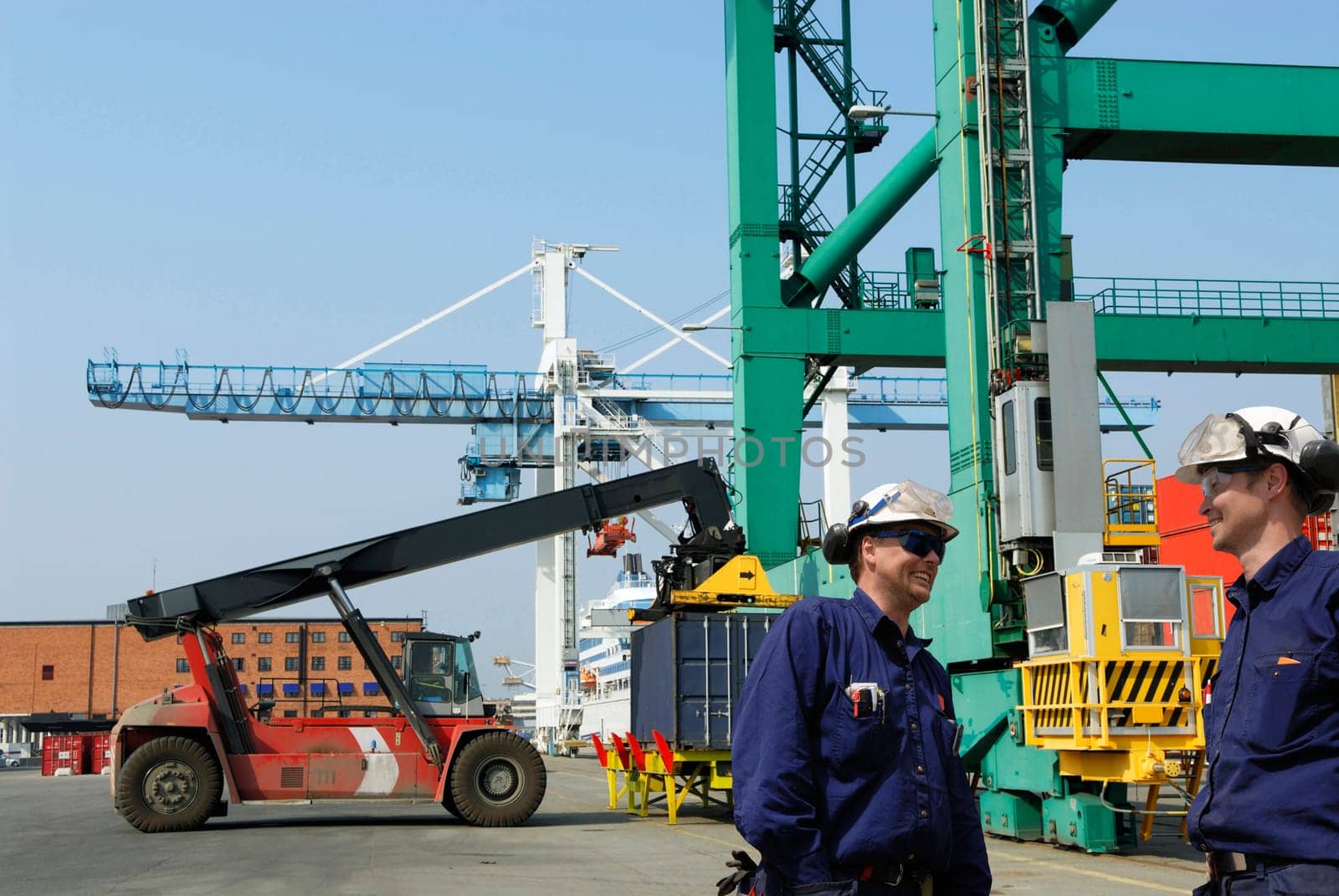 busy container-port, trucks stacking containers from crane, two port-workers in foreground