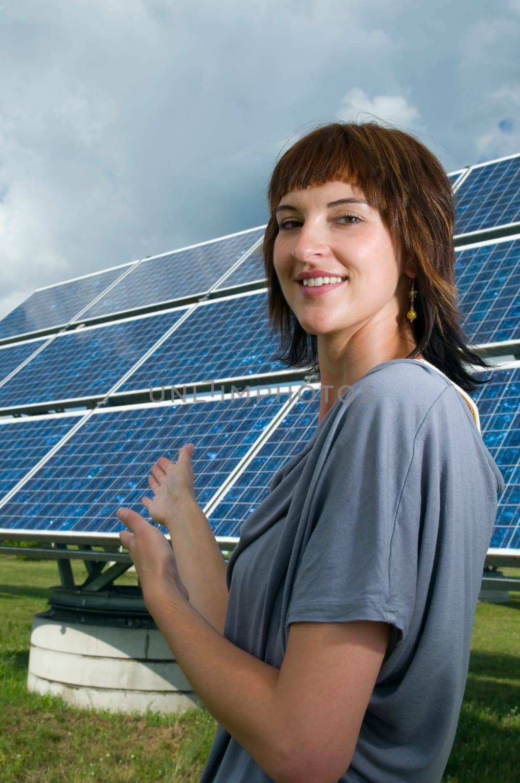 Upper body shot of a young brunette woman in casual casual dress on a green meadow in summer pointing to a photovoltaic system behind her