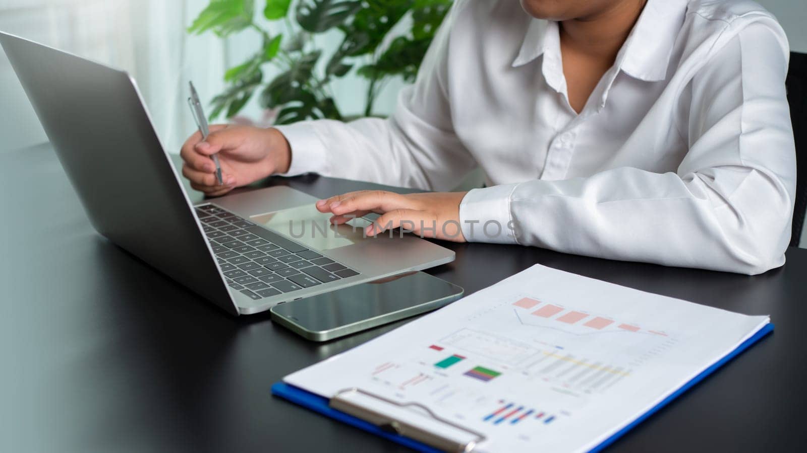 Businesswoman holding a pen and using a computer to work sits in a modern office with copy space. by Unimages2527