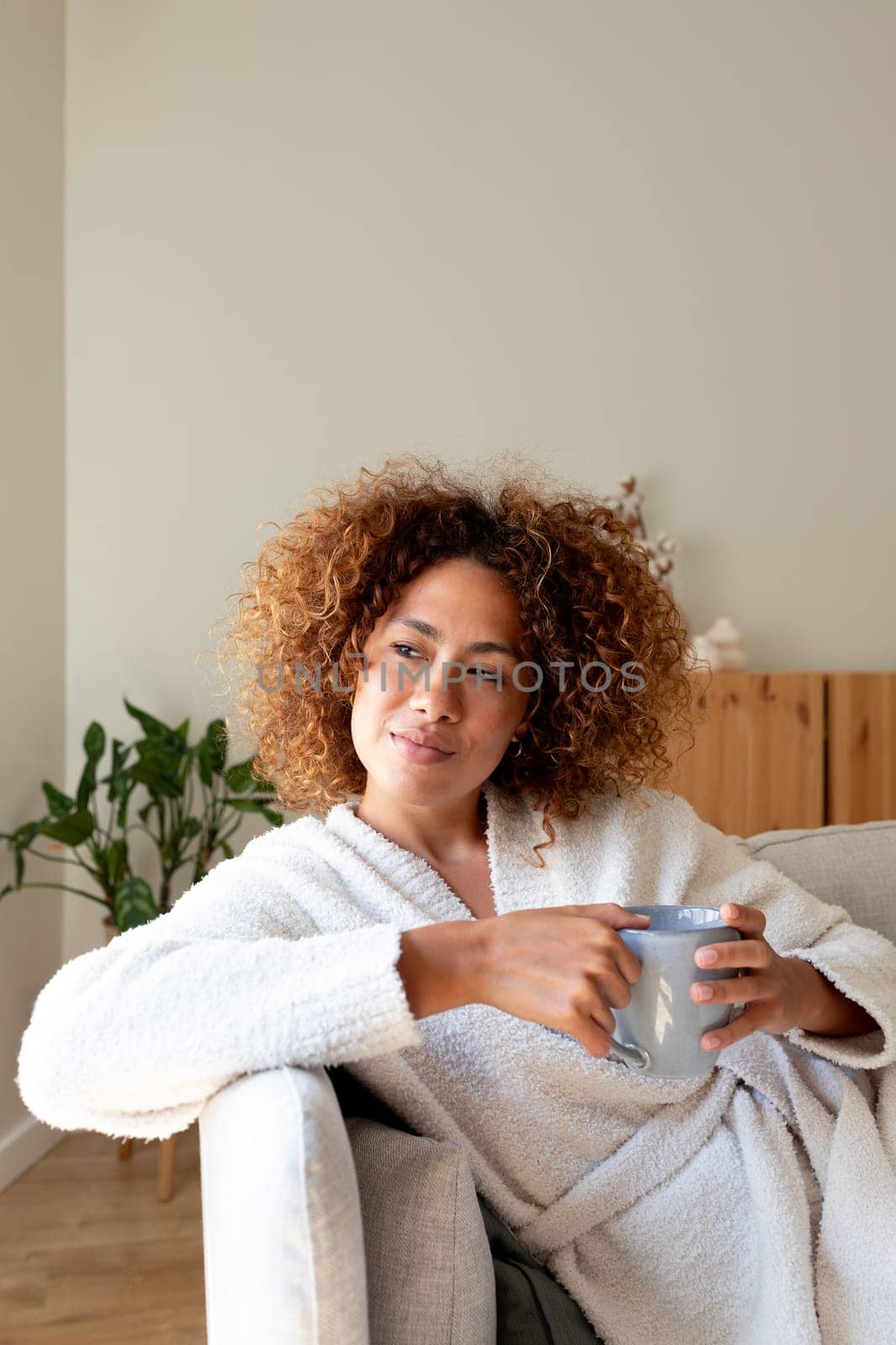Pensive multiracial woman relaxing at home, sitting on the sofa drinking tea looking out the window. Copy space. Vertical image. Lifestyle concept.