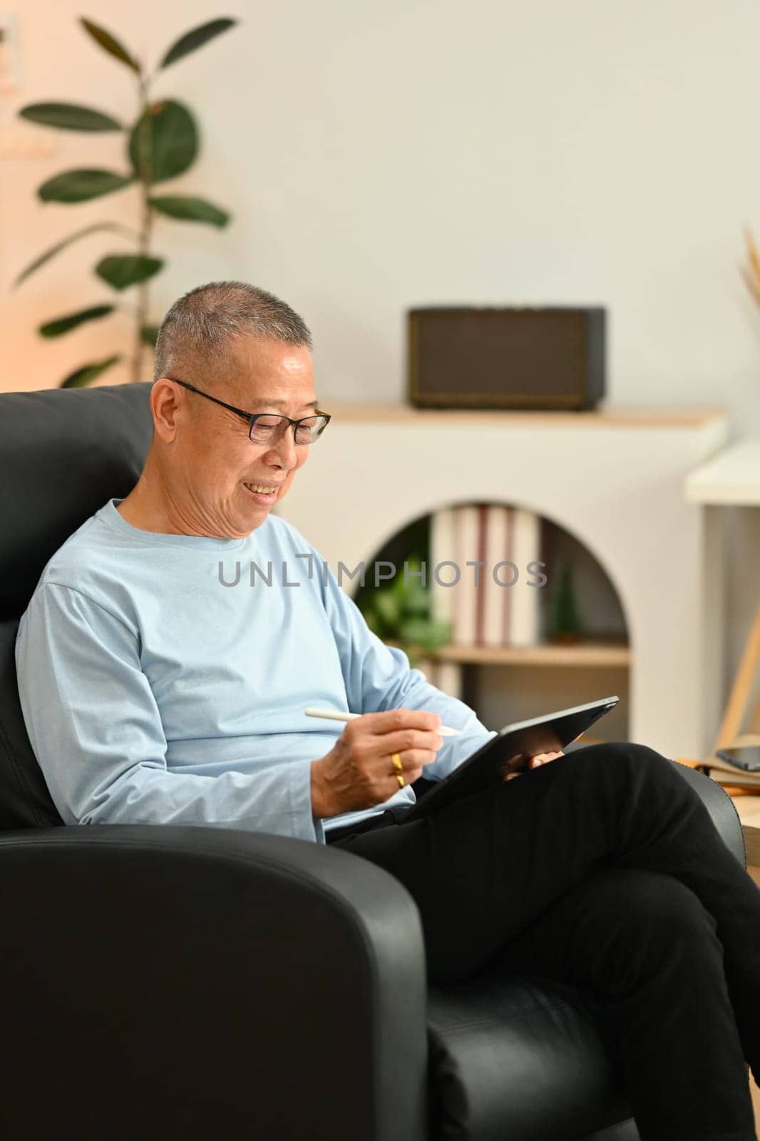 Smiling senior man in glasses using digital tablet while sitting on armchair at home.
