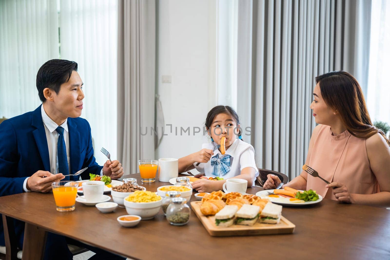 Asian family father, mother with children daughter eating healthy breakfast food on dining table kitchen in mornings together at home before father left for work, happy couple adult family concept