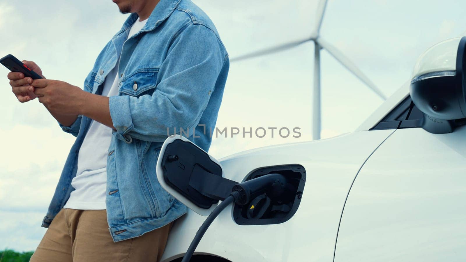 Progressive man with his electric car, EV car recharging energy from charging station on green field with wind turbine as concept of future sustainable energy. Electric vehicle with energy generator.