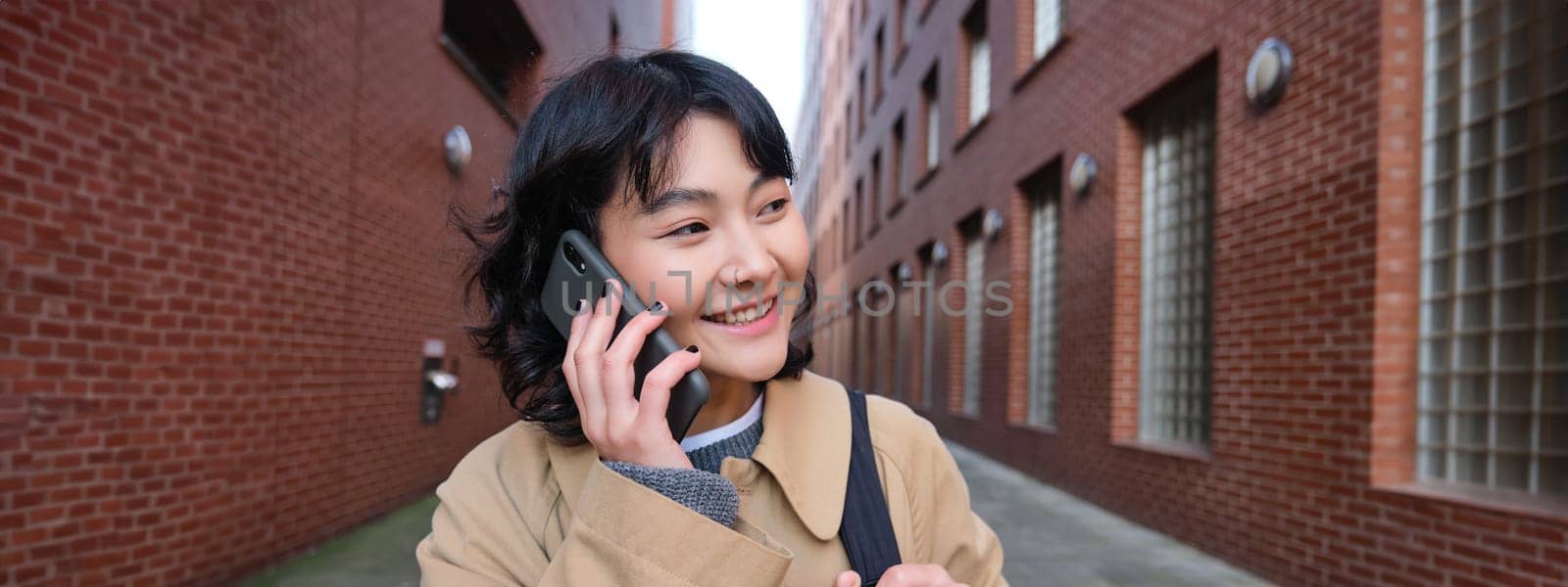 Portrait of young korean woman walking down street with phone, talking with someone, makes a call, has telephone conversation.