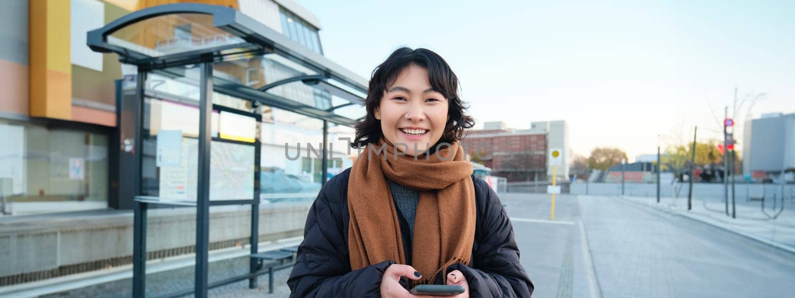 Portrait of Korean woman in winter jacket, standing with smartphone, waiting for bus on stop, looking at mobile phone app checking public transport application by Benzoix