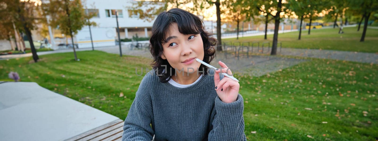 Portrait of asian woman, student in park, sits on bench with digital tablet and a pen, thinking, looking aside thoughtful, making notes, does her homework outdoors.