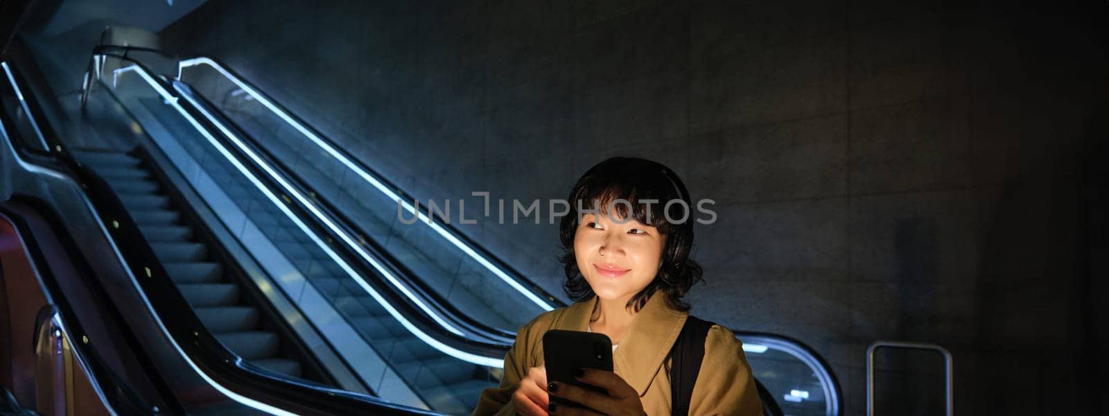 Smiling Korean girl in headphones, listens music, uses mobile phone in tube, stands near escalator in dark, looks cheerful.
