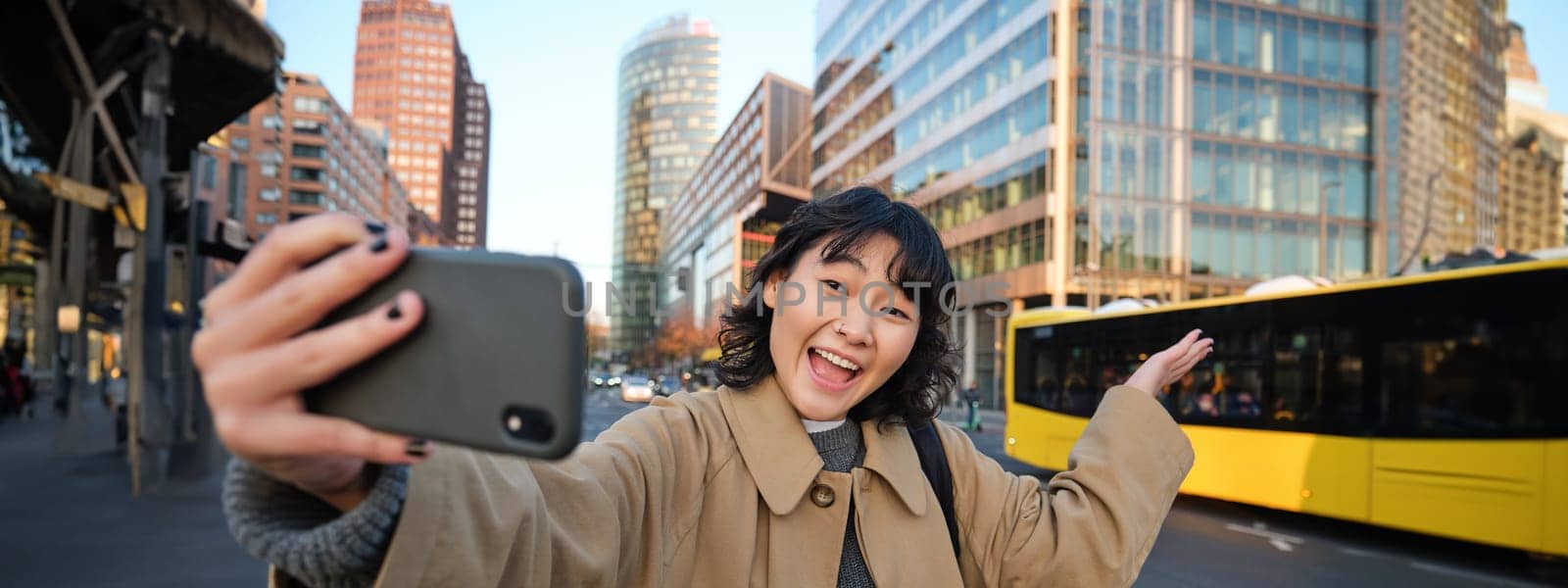 Upbeat asian girl takes selfie with smartphone in city centre, showing something behind her with smiling face. Tourist goes sightseeing.