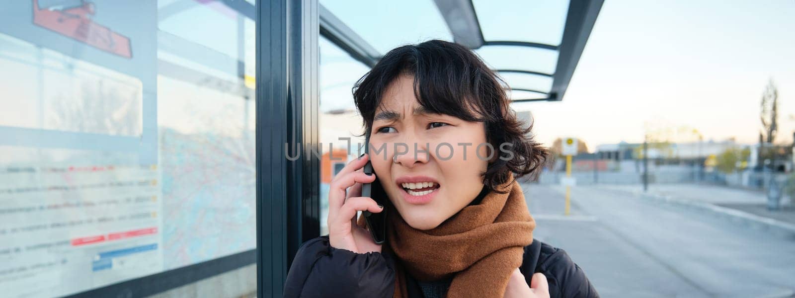 Young woman looks frustrated and disappointed, talks on mobile phone, stands on bus stop, receives bad news. Copy space
