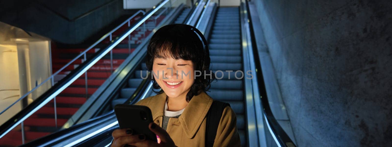 Portrait of girl student in headphones, listens music, commutes, goes down escalator, looks at smartphone with pleased smile, happy face.