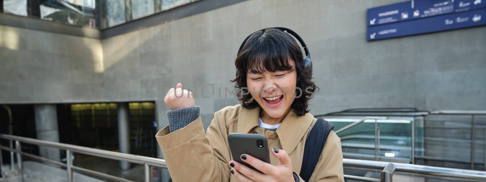 Happy asian girl in headphones, looking at mobile phone and celebrating, reading message with good news, screams yes with excitement, stands on street.