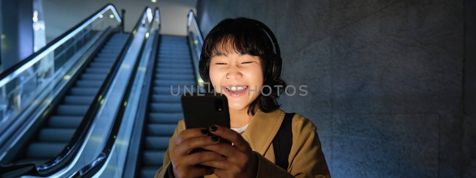 Laughing cute Korean girl on escalator, going down to tube, underground or metro, using mobile phone, watching smth funny on smartphone app by Benzoix