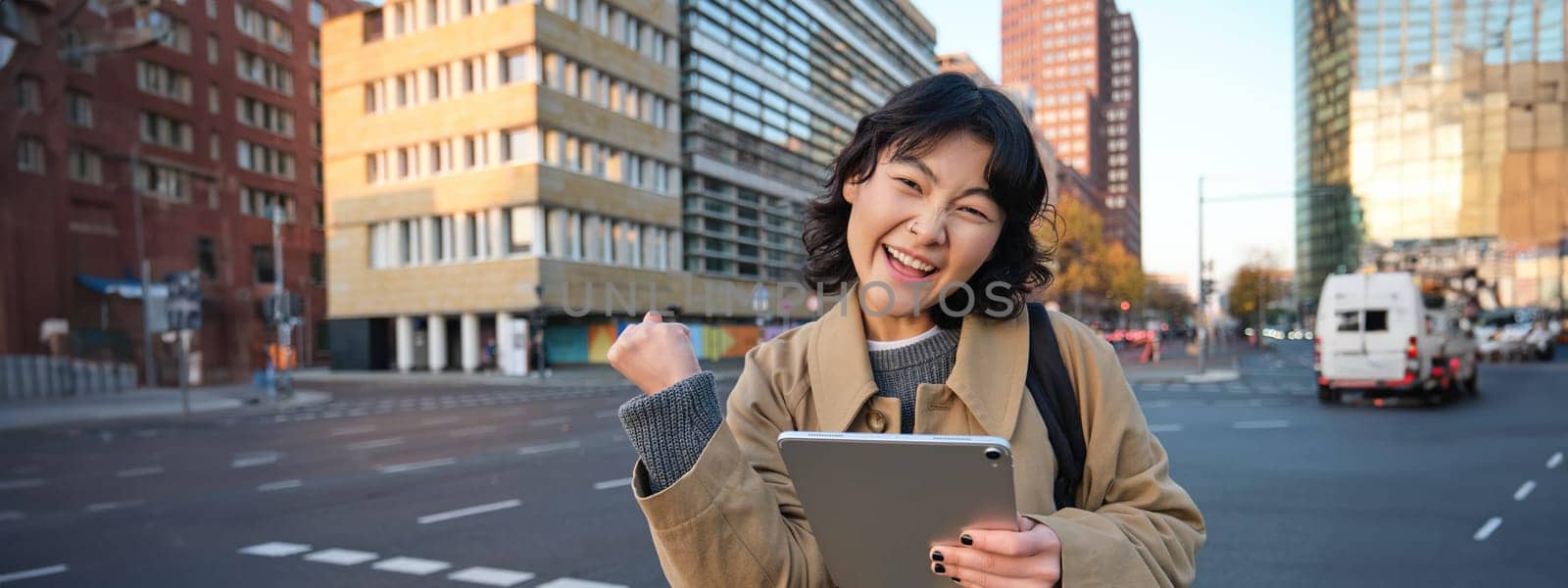 Enthusiastic asian girl with digital tablet, smiling and looking amazed, happy with something good, holding digital tablet, standing on street by Benzoix