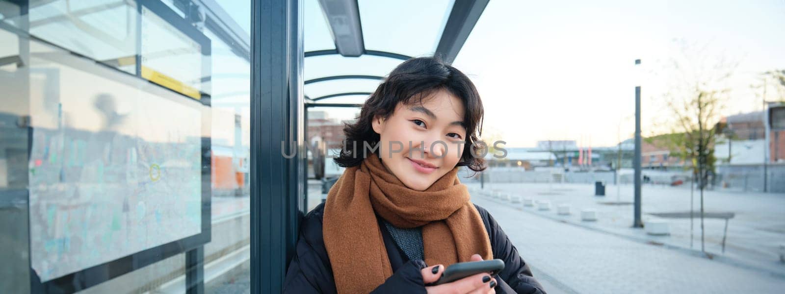 Beautiful young woman, standing on bus stop, waiting for her public transport, using mobile phone, using smartphone application by Benzoix