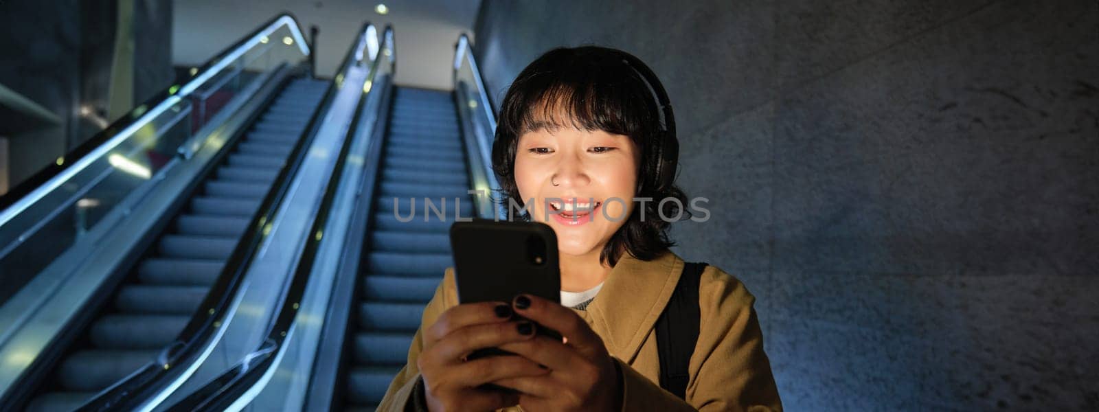 Stylish young girl, student in headphones, listening music on her smartphone, reading news on phone while going down on escalator.
