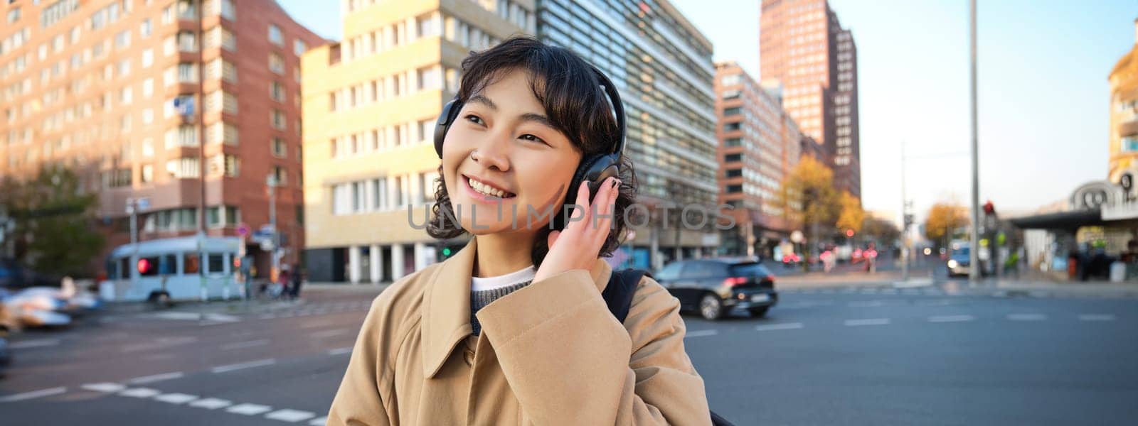 Portrait of young smiling korean girl, walking along city centre, listening music in headphones and holding mobile phone by Benzoix