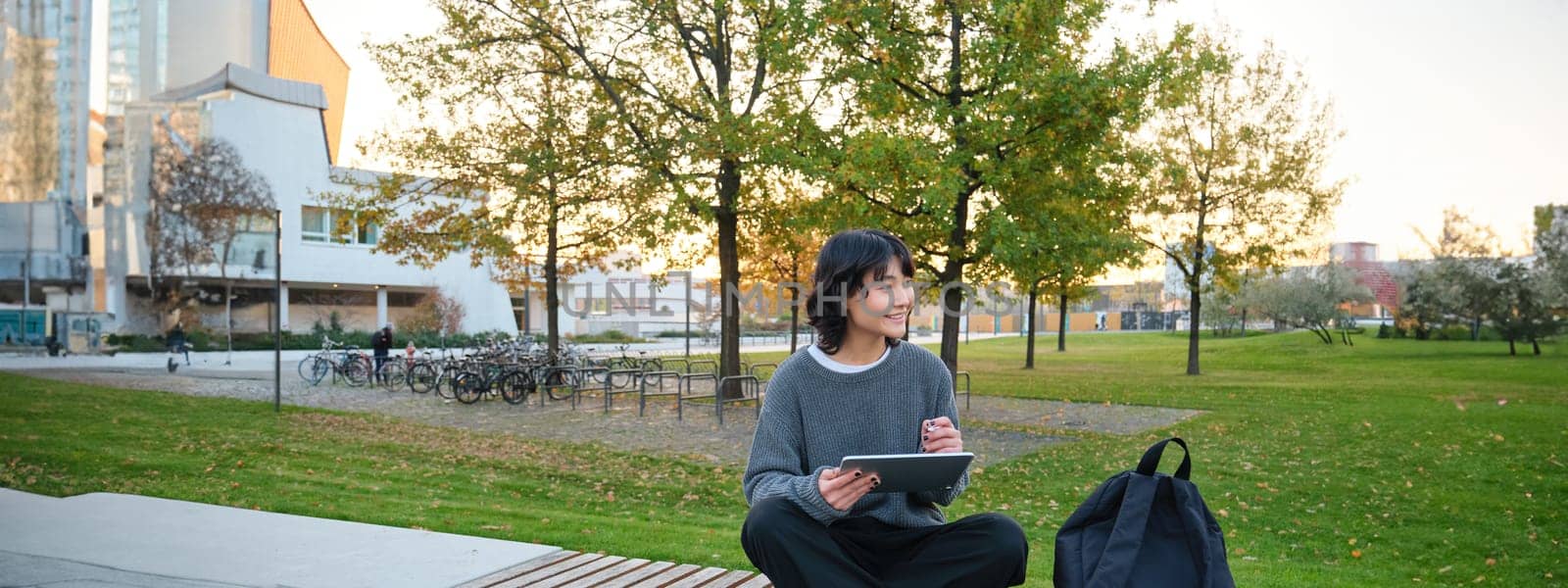 Young girl, korean artist or art student sits in park with digital tablet, draws with graphic pen, scatches a design or project, looks around by Benzoix