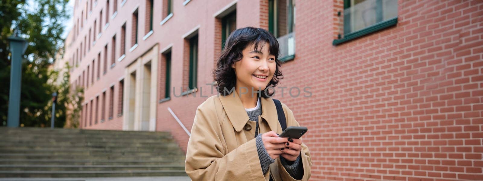 Stylish korean hipster girl, walks in city centre, sends text message, uses location map application on smartphone while walking down the street, wears trench coat.