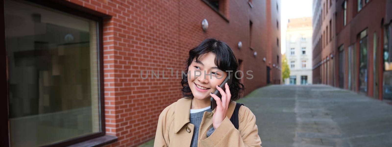 Portrait of young korean woman walking down street with phone, talking with someone, makes a call, has telephone conversation.