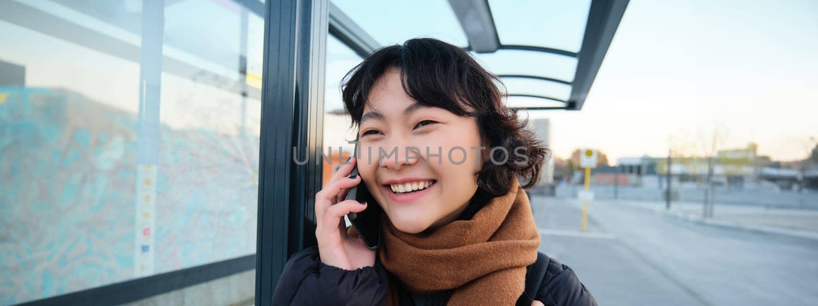 Close up of cute Korean woman, making a phone call, talking and smiling on telephone, standing in winter jacket on bus stop, waiting for her transport to arrive by Benzoix