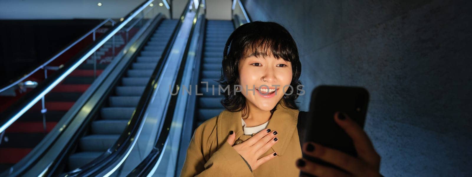 Portrait of asian girl uses smartphone, listens music in headphones, goes down escalator in city centre in evening, looking surprised at phone, gasping amazed.