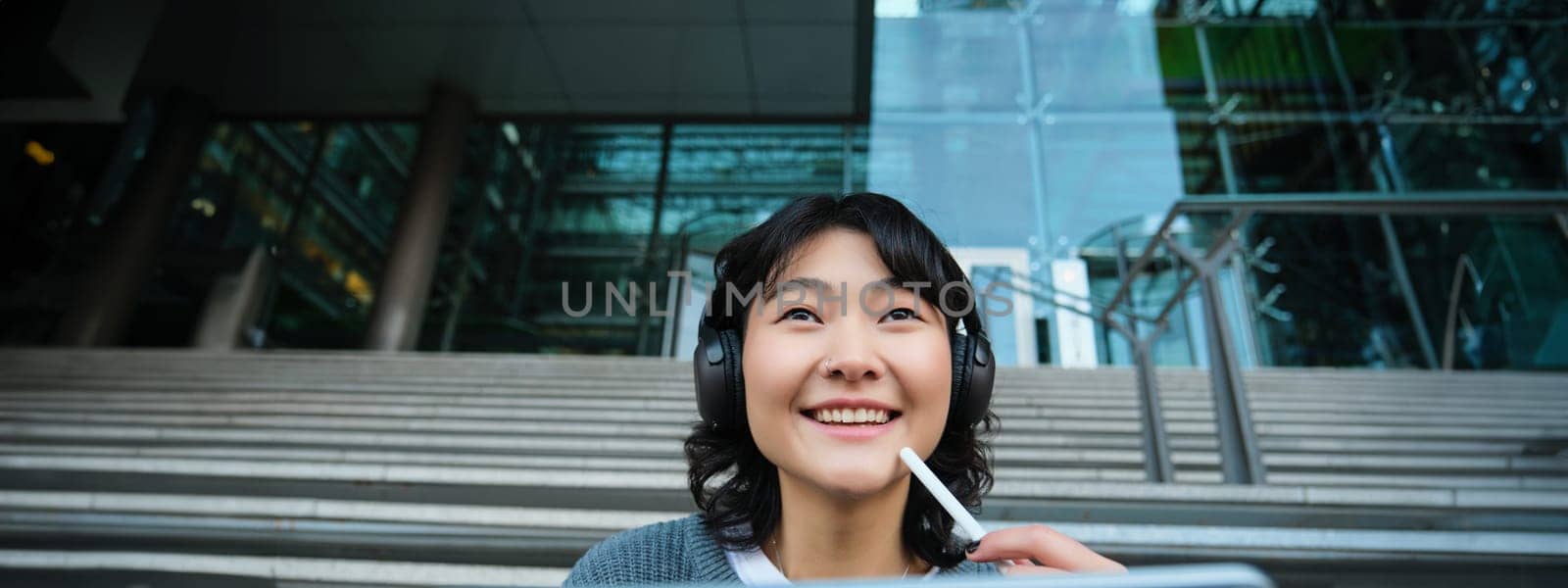 Close up of smiling asian girl feels inspired while draws on digital tablet, makes graphic design project, pencils user interface assignment, sits on stairs near campus by Benzoix