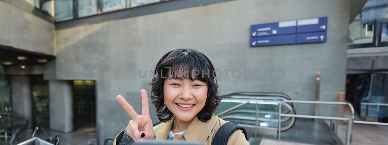 Cute and stylish Korean girl, wears headphones, takes selfie on smartphone, tourist records video or makes a photo, stands on street.