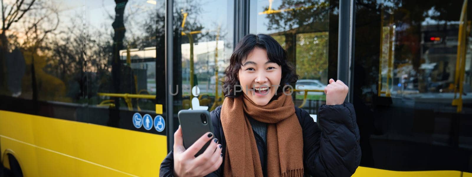 Enthusiastic asian woman, standing on bus stop with smartphone, looking at phone screen with amazed, triumphing face, winning, hear great news on video chat by Benzoix