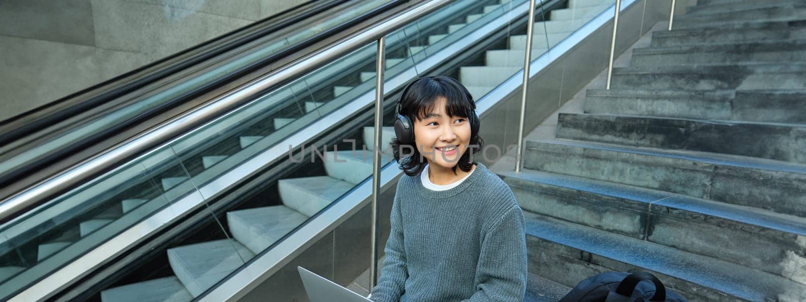 Portrait of beautiful young korean woman, student sits on stairs in public place, listens music in headphones, works on project on laptop, works remotely by Benzoix