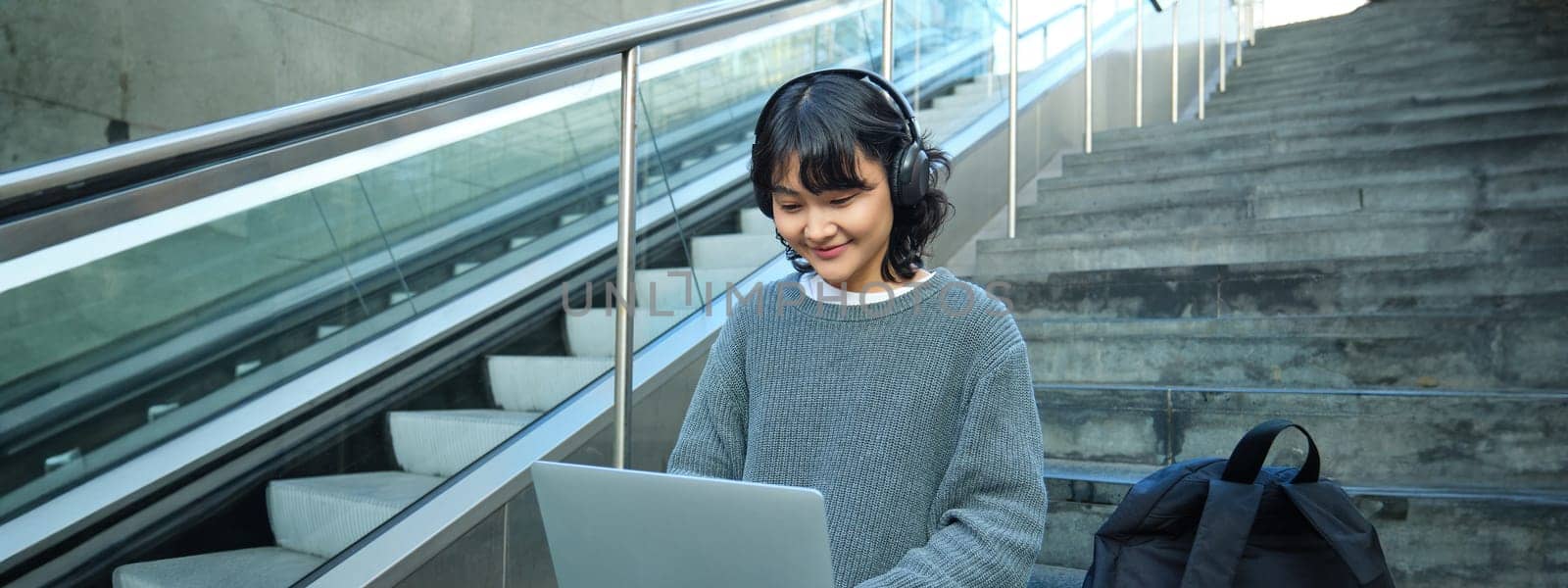 Portrait of beautiful young korean woman, student sits on stairs in public place, listens music in headphones, works on project on laptop, works remotely by Benzoix